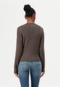 Brauner langärmeliger Strickpullover mit einer V-förmigen Naht auf der Rückseite, kombiniert mit auffälligen blauen Jeans aus Denim mit Gesäßtaschen.