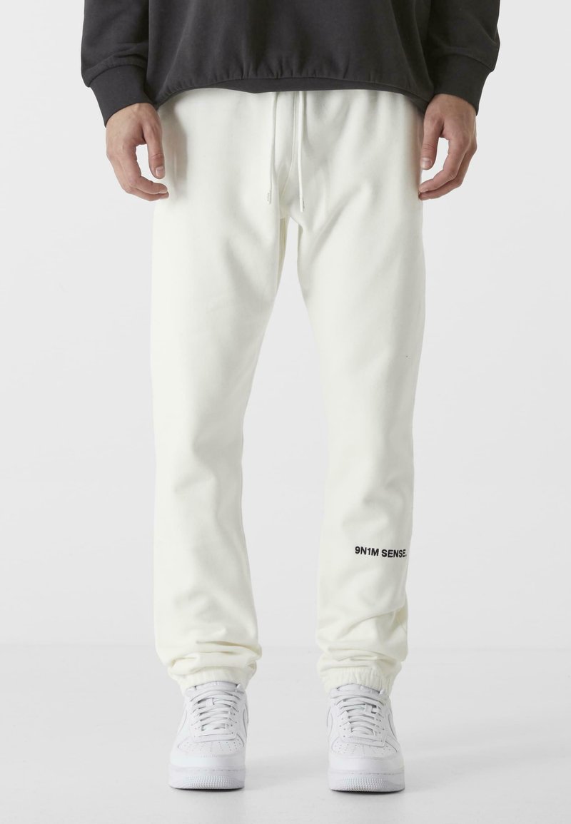 White sweatpants made of soft fabric, featuring an elastic waistband with drawstring, ribbed cuffs, and a black logo detail on the left leg.