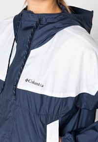 Granatowo-biała kurtka Columbia z kapturem, regulowanymi ściągaczami i kieszenią na piersi z logo. Wykonana z lekkiego materiału.