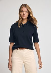 Granatowa koszulka polo z klasycznym kołnierzykiem, krótkimi rękawami, z trzema guzikami i luźnym krojem, w zestawieniu z beżowymi spodniami i brązowym paskiem.