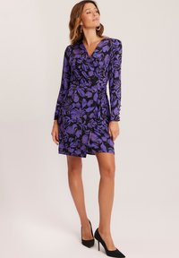 Fioletowa sukienka w kwiaty, z długimi rękawami i dekoltem w serek. Posiada czarne tło oraz detal w postaci boku z związaniem. Noszona z czarnymi szpilkami.