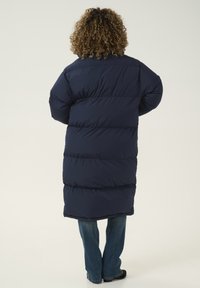 Abrigo tipo puffer azul marino con secciones acolchadas en horizontal, mangas largas y un dobladillo ligeramente curvado, combinado con unos vaqueros azules y zapatos negros.
