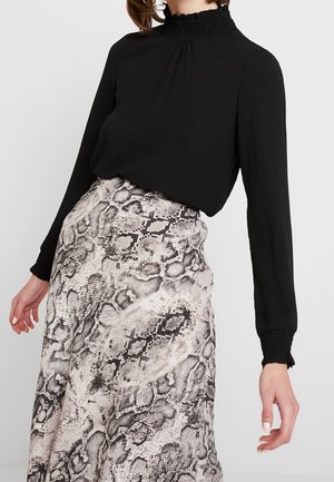 Vrouw draagt een zwarte top met lange mouwen met een hoge, geplooide kraag en een witte en grijze rok met slangenprint.