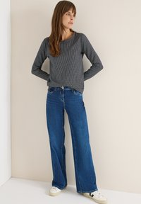 Langärmliges graues Oberteil mit strukturiertem Muster, kombiniert mit weit geschnittenen blauen Jeans. Weiße Sneakers mit schwarzen Akzenten runden den Look ab.