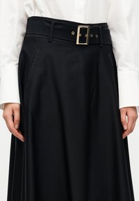 Μαύρη φούστα με φαρδιά ζώνη μέσης που διαθέτει ορθογώνιο χρυσό αγκράφα και θηλιές για τη ζώνη. Το υλικό είναι λείο, με πλαϊνές τσέπες και αέρινο σχέδιο.