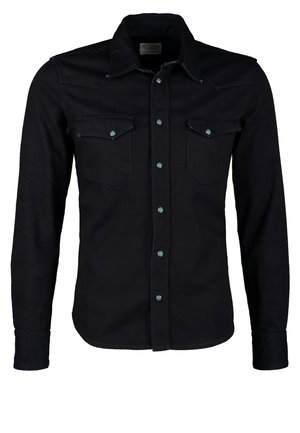 Hombre con chaqueta negra, camisa gris y pantalones cortos caqui, sosteniendo una bolsa de viaje marrón y beige en la mano derecha, mano izquierda en el bolsillo.