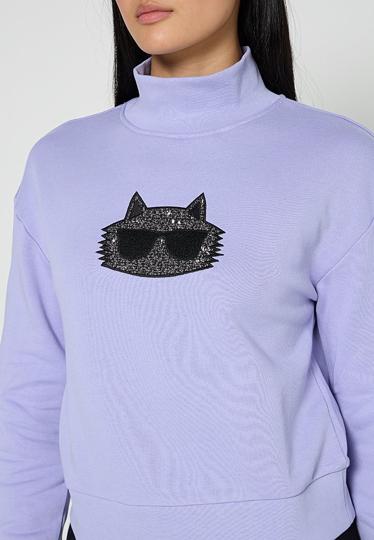 Sudadera lila con un cuello alto que presenta una cara de gato negra y brillante con gafas de sol como elemento central del diseño. Tela suave y texturizada.