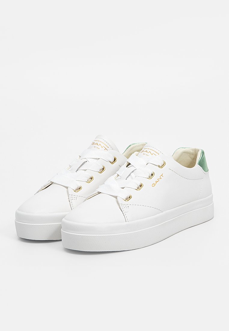 Weiße Sneakers aus Leder mit goldenen Ösen, flachen Schnürsenkeln und einem grünen Akzent an der Ferse. Abgerundete Spitze und strukturierte weiße Sohle.