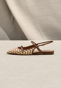 Κωνικό καφέ και λευκό πλεκτό επίπεδο σανδάλι με λεπτά καφέ λουριά αστραγάλου και χαμηλό τακούνι σε ουδέτερο φόντο.