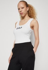 White ribbed tank top featuring "GAP" logo in black, paired with black elastic waist pants with pockets. Smooth fabric texture.