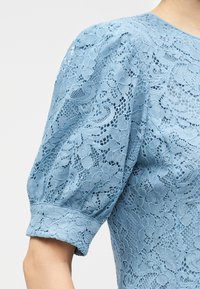 Κοντινό πλάνο ενός ανοιχτό μπλε δαντελένιου μπλουζιού με λουλουδένια σχέδια και φουσκωτά κοντά μανίκια που τελειώνουν σε εφαρμοστό μανσέτα.