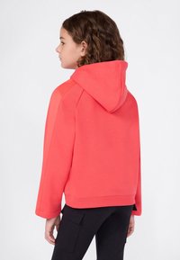 Dziewczyna z kręconymi włosami w czerwonym hoodie i czarnych spodniach cargo, stojąca i zwrócona w lewo na jednolitym jasnym tle.