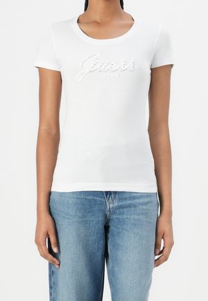 Kobieta w dopasowanym białym t-shircie z krótkim rękawem z logo "Guess" z cyrkoniami i jasnoniebieskich dżinsach na jednolitym tle.