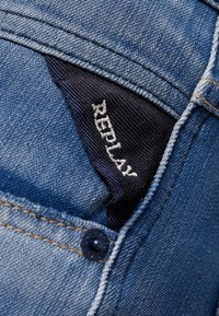 Tasca in denim blu con un'etichetta in tessuto blu scuro con la scritta "REPLAY" in lettere bianche, accentuata da un bottone visibile e da cuciture gialle.