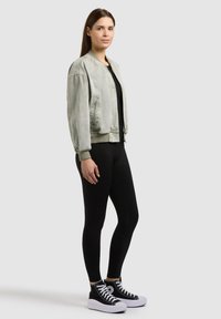 Femme debout, légèrement tournée vers la droite, portant une veste bomber gris clair, des leggings noirs et des baskets montantes noires avec des semelles blanches.