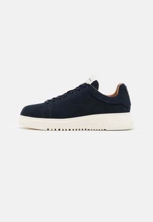Lavtopet mørkeblå sneakers med hvid tyk sål og sorte snørebånd, vist fra siden på en lys grå baggrund.