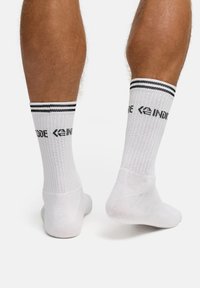 Białe skarpetki sportowe z czarnymi paskami u góry, prążkowaną fakturą i nadrukowanym logo po bokach. Standardowa długość do kostki.