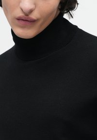 Close-up de uma pessoa a usar um suéter de gola alta preto com nervuras, focando na parte inferior do rosto e no torso superior contra um fundo simples.
