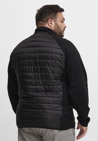 Black fleece jacket with a padded back; features a high collar, long sleeves, and a quilted texture in matte black material.