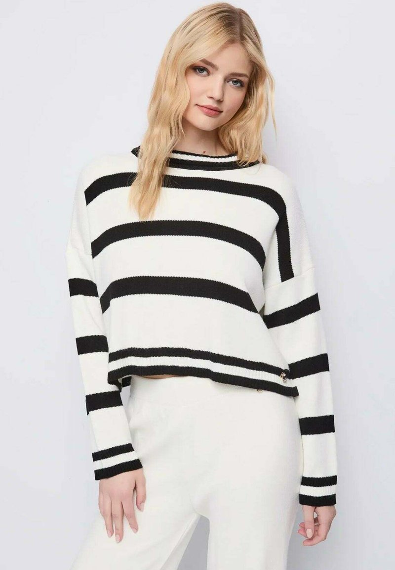 Donna bionda con un maglione bianco a larghe strisce orizzontali nere e pantaloni bianchi, in piedi contro uno sfondo chiaro e semplice.