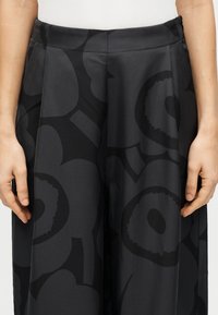 Μαύρο παντελόνι με φαρδιά φάσα που διαθέτει ένα λουλουδάτο μοτίβο σε ελαφρώς πιο σκούρα απόχρωση. Ομαλή υφή με ραμμένη ζώνη στη μέση και πιετά σχεδίαση.