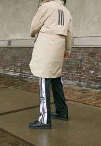 Beżowa parka o kroju asymetrycznym, z trzema pionowymi paskami z tyłu, w połączeniu z czarnymi i białymi spodniami dresowymi na napy oraz czarnymi sneakersami.