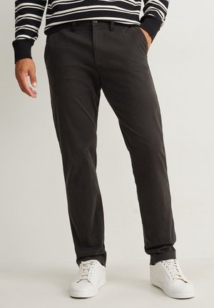 Mann trägt schmale schwarze Hose, weiße Sneakers und ein schwarz-weiß gestreiftes langärmeliges Oberteil, steht mit einer Hand in der Tasche.