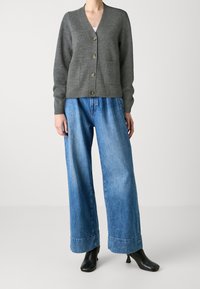 Grauer Wollcardigan mit V-Ausschnitt, sechs Knöpfen und vorderen Taschen, kombiniert mit lässigen, hellblauen Jeans mit weitem Bein und schwarzen Schuhen.