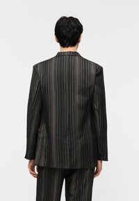 Άνδρας που στέκεται με το πρόσωπο προς τα πίσω, φορώντας μαύρο κοστούμι με λεπτές κάθετες χρυσές ρίγες στο σακάκι και το παντελόνι, μπροστά σε απλό φόντο.