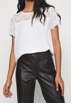 Vrouw draagt een witte korte mouwen top van kant en geplooide stof, gecombineerd met zwarte leren broek met hoge taille.