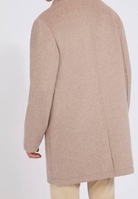 Mand iført en beige uldfrakke med lange ærmer, set fra baggrunden mod en ensfarvet lys baggrund.