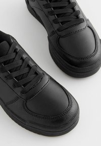 Baskets de sport noires avec une tige en cuir lisse, un col rembourré et des lacets plats. Accents texturés notables sur le côté et semelle en caoutchouc.