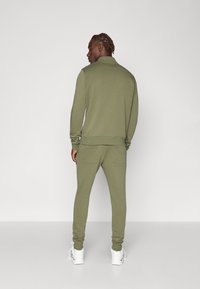Olivengrønn treningsdrakt med glidelåskrage, tettsittende mansjetter og to baklommer. Stoffet virker mykt med en glatt tekstur. Hvite joggesko.