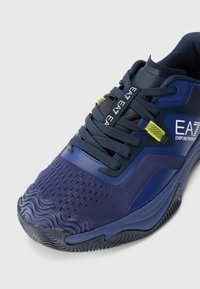 Navy sneaker featuring a textured upper, visible ventilation holes, and a rubber sole with a wavy pattern. Accents include yellow tags.