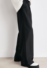 Schwarze weite Hosen aus glattem Stoff, mit hoher Taille und gefaltetem Design. Kombiniert mit klobigen Schuhen mit weißer Sohle.
