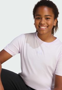 Jasnoróżowa koszulka z krótkim rękawem z subtelnym logo w formie trzech pasków na piersi. Miękka tkanina z okrągłym dekoltem, zestawiona z ciemnymi spodniami.