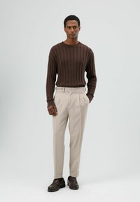 Bruine geribbelde trui, beige op maat gemaakte broek met plooien en knoopdetails, gecombineerd met donkerbruine schoenen. Neutrale achtergrond.