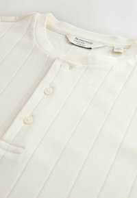 Jasno-beżowy, prążkowany sweter z okrągłym dekoltem, trzema guzikami i małą kieszonką na piersi. Gładka tekstura i dopasowany krój.