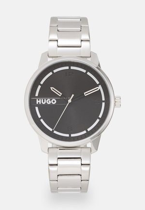 Montre-bracelet en métal argenté avec un cadran noir, des index horaires blancs et le logo "HUGO". Elle possède un bracelet en acier inoxydable et un boîtier rond lisse.