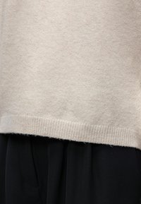 Gros plan sur l'ourlet d'un pull beige tricoté sur un tissu noir plissé, montrant la texture et la superposition des vêtements.