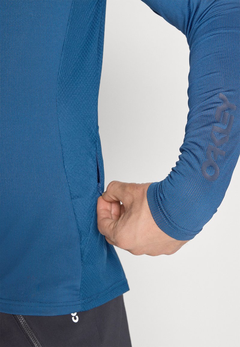 Camisa deportiva azul de manga larga con tejido texturizado y bolsillo lateral. Cuenta con un logo en la manga y un ajuste ceñido alrededor de la muñeca.