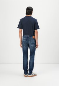 Marineblå bomulds t-shirt, blå denim jeans med fem lommer og læderlap, afslappede hvide sneakers, set bagfra mod en neutral baggrund.