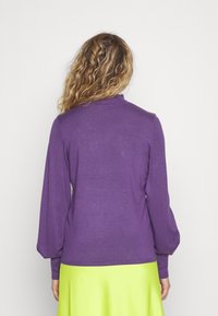 Fialový svetr s dlouhými, nadýchanými rukávy a ribbovaným límcem, kombinovaný s jasně zelenou sukní. Měkká textura, přiléhavý střih.