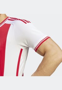 Prawa ręka zgięta, ubrana w biało-czerwoną sportową koszulkę z krótkim rękawem i trzema czerwonymi paskami na ramieniu.