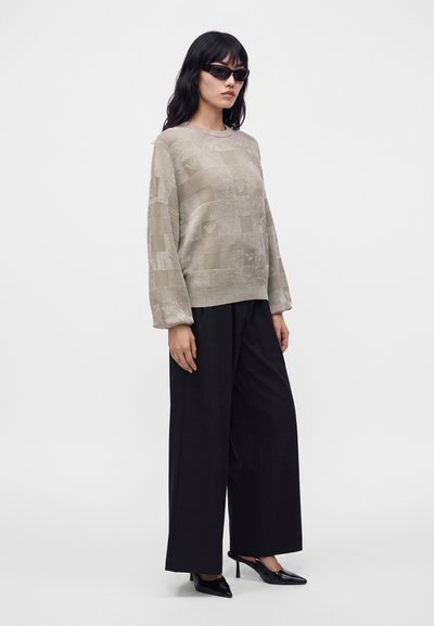Femme portant un pull beige à motifs, un pantalon noir large, des talons noirs et des lunettes de soleil foncées, debout devant un fond uni.