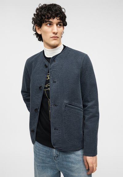 Μπλε σκούρο, υφασμάτινο σακάκι με στρογγυλή γιακά, κλείσιμο με κουμπιά και πλαϊνή τσέπη. Φορεμένο πάνω από μαύρο πουκάμισο και συνδυασμένο με ανοιχτό μπλε τζιν.