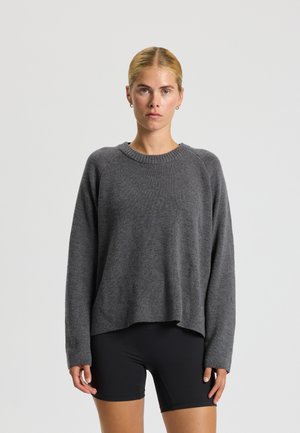 Giovane donna con capelli biondi, indossa un maglione grigio a maglia e pantaloncini neri, in piedi su uno sfondo chiaro uniforme.