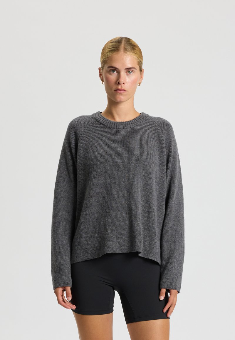 Jeune femme aux cheveux blonds portant un pull en maille gris et un short noir, debout devant un fond clair uni.