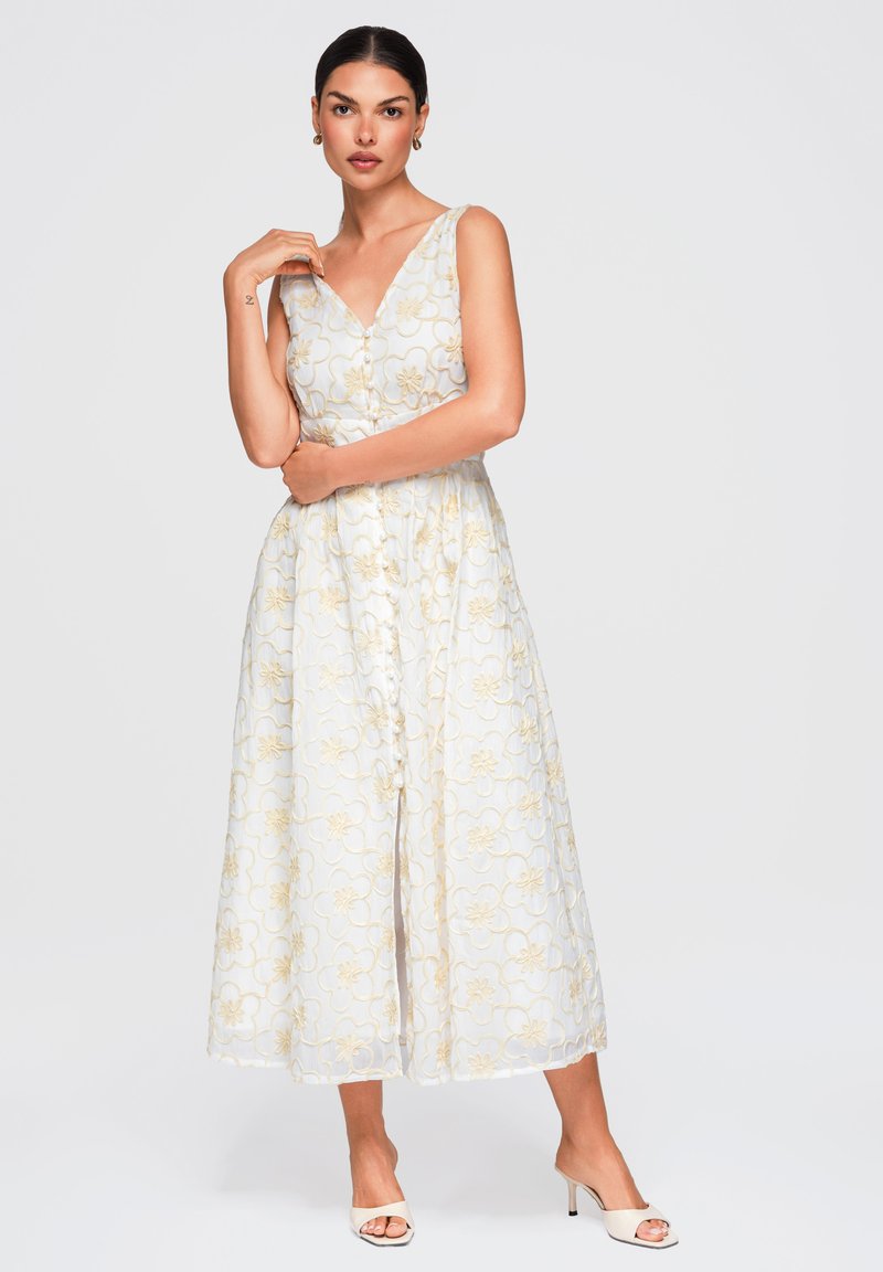 Femme debout portant une robe midi blanche sans manches avec une broderie florale jaune et des sandales blanches à talons ouverts sur un fond uni.