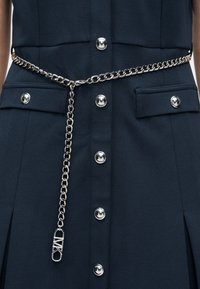 Μπλε φόρεμα ναυτικού στιλ με ασημένιες λεπτομέρειες κουμπιών και ασορτί ασημένια ζώνη με αλυσίδα που φέρει ένα μοτίφ με λογότυπο να κρέμεται μπροστά.